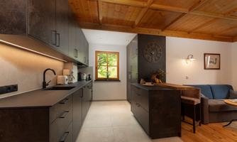 A modern kitchen with dark furniture and a wooden ceiling. Daylight streams in through the window, and there is a cozy seating area.
