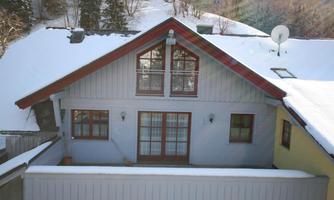 A modern house with a red roof, surrounded by snow and trees. It has large windows and a terrace.