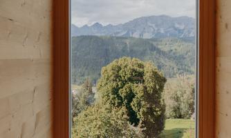 A large window frame reveals a green landscape with a tree and mountains in the background. The sky is cloudy, giving the scene a calm atmosphere.