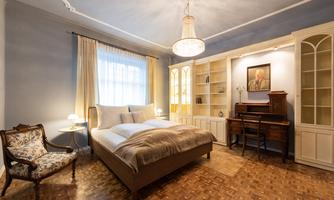 A cozy bedroom with a double bed, an old desk, and a comfortable armchair. The room is brightly lit and has large windows with curtains.