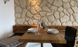 A cozy dining area with a wooden table and modern chairs. The wall is clad in natural stones, and glasses and plates are set.