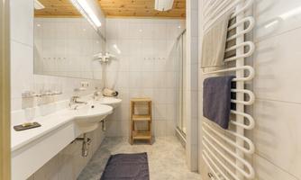 A modern bathroom with white tiles and a wood ceiling. It is equipped with a sink, a shower, and a towel holder.