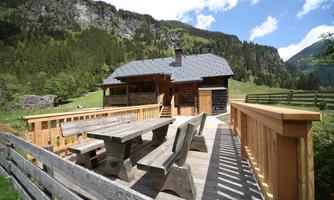 A cozy cabin in the mountains with a large terrace and wooden furniture. In the background, green meadows and tall mountains can be seen.