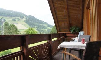 A cozy balcony with wooden furniture and a table. In the background, green hills and trees can be seen.