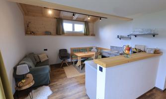 A cozy living area with a sofa and a wooden table. The walls are light, and large windows provide plenty of daylight.