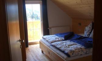 A cozy bedroom with a double bed and wooden floor. A window overlooking a balcony and daylight illuminating the room.
