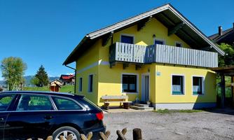 A yellow house with a balcony and a green surrounding. In front of the house, there is a black car.