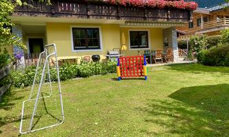 A beautiful garden with grass and a playground. In the background, there is a cozy house with a flower balcony.