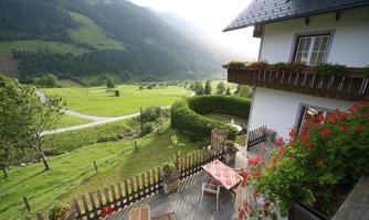 Insight into a picturesque landscape with a green hill and gentle mountains. On the terrace, there are tables and chairs, surrounded by blooming plants.
