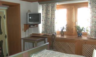 A cozy room with a bed, a table, and a television. The windows have beautiful curtains and let in plenty of daylight.