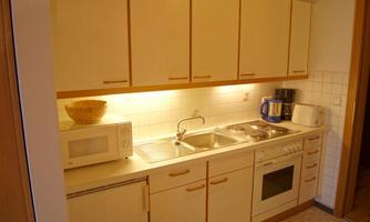 A modern kitchen with light wooden furniture and a countertop. It is well-equipped with a microwave, a kettle, and a sink.