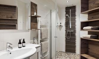 A modern bathroom with a walk-in shower and wooden shelves. The walls are painted in neutral colors and there are personal care products on the sink.
