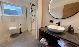 A modern bathroom with a large shower, a round mirror, and an elegant sink. Fresh towels are ready on the shelf.