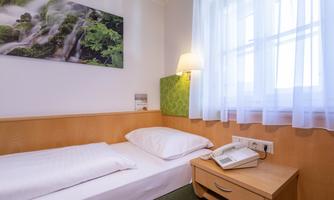 A simple single room with a bed, a bedside table, and a telephone. A green landscape picture hangs on the wall.