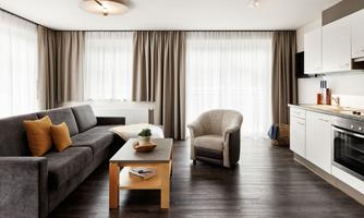 Modern living room with a gray sofa and an armchair. Large windows let in plenty of daylight and there is a bright, practical kitchenette.