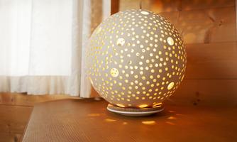 An elegant spherical lamp with many small holes that emits warm light. It stands on a wooden table next to a window with curtains.