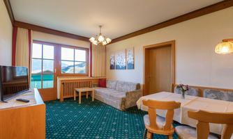A cozy living room with a sofa, a table, and chairs. The room has large windows overlooking the landscape.