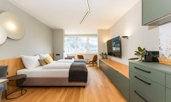 A modern room with two beds and large windows. The decor is minimalist and elegant, with wood and green tones.