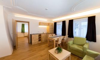 A modern living room with a green armchair, a wooden table, and an open kitchen. Bright walls and large windows provide plenty of daylight.