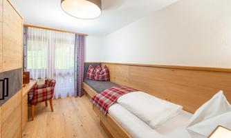 A cozy bedroom with wooden paneling and a single bed. The room has large windows with curtains and a rustic seating area.