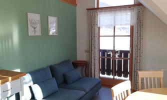 A cozy living room with a blue sofa, a table, and beautiful pictures on the wall. The windows provide plenty of daylight and offer a view outside.