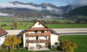 A large house with several balconies, surrounded by green meadows and mountains. In the background, the peaks of the mountains are visible with some clouds.
