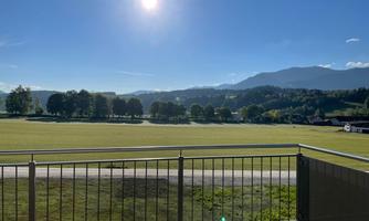 A sunny view from a balcony overlooking a vast meadow landscape with trees in the background. The mountains stretch across the horizon under a clear blue sky.