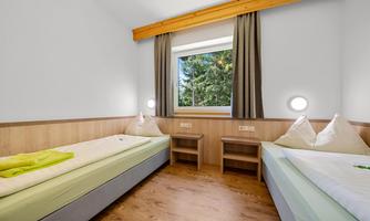 A cozy room with two single beds and wooden paneling. The window offers a view of greenery and provides plenty of natural light.