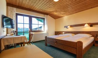 A cozy bedroom with wooden decor, two beds, and a television. There is a table with chairs and large windows that let in plenty of daylight.