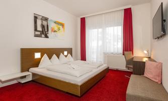 A modern bedroom with a large double bed and red carpet. The walls are light, and there are large windows with red curtains.