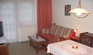 A cozy living room with a red sofa and a wooden table. Large windows let in plenty of light, and the decor is simple and inviting.