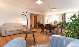 A modern living room with comfortable furniture, a round table, and a cozy atmosphere. The large windows provide plenty of daylight.
