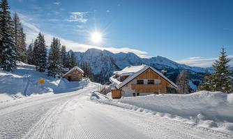 A picturesque winter landscape with snow-covered houses and majestic mountains in the background. The sun shines in the clear blue sky.