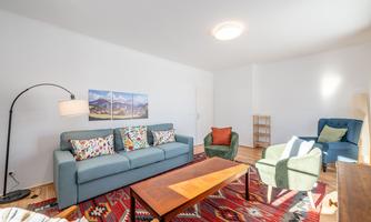 A cozy living room with a blue sofa and colorful cushions. The room has a modern rug and a bright, friendly atmosphere.