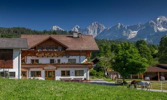 A picturesque house in the mountains, surrounded by green meadows and trees. Impressive mountain peaks can be seen in the background.