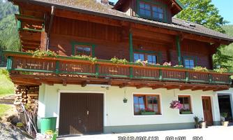 A traditional wooden house with a balcony adorned with flowers. In the foreground, there are stacks of wood and a parking lot.