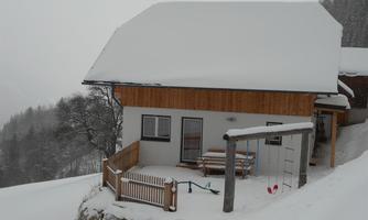 A house in the snow with a wooden porch and surrounding trees. The landscape appears calm and wintry.