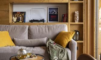 A cozy living room with a bright sofa and a round table. Wooden shelves and large windows create a warm atmosphere.