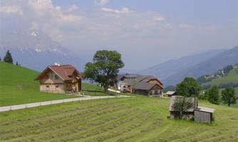 A picturesque landscape with two wooden houses and a large tree. In the background, gentle hills and mountains rise beneath a clear sky.