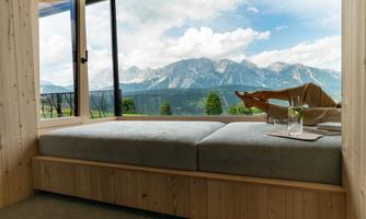 A cozy room with a large window front that shows a stunning mountain landscape. The surroundings are bright and inviting, perfect for relaxing.