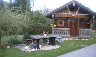 A charming wooden cottage with an inviting porch and colorful flowers. In front stands a rustic stone table in a green setting.
