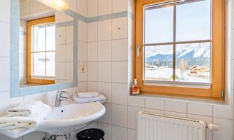 A bright bathroom with windows offering a view of the mountains. The walls are tiled, and there is a sink as well as towels.