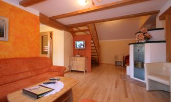 A cozy living room with an orange sofa and a wooden table. In the background, a staircase and a television can be seen.