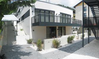A modern building with large windows and balconies. The outdoor area is designed with gravel and plants, surrounded by stairs and other buildings.