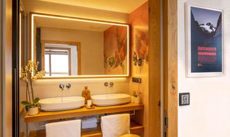 A modern bathroom with two sinks and a lit mirror. The furnishings are made of wood, and there is a decorative plant on the sink.