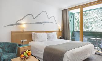 A modern hotel room with a double bed and a cozy seating area. Large windows provide plenty of light and access to a balcony.