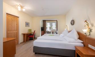 A cozy room with a double bed, a table, and a chair. Large windows bring plenty of daylight into the room.