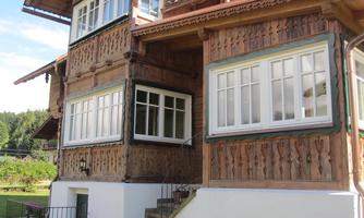 A traditional wooden house with detailed carvings and large windows. The surroundings are green and inviting.