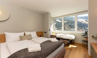 A modern bedroom with a large window that offers a view of the mountains. There is a double bed and a relaxing lounge chair.