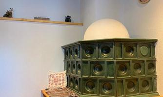 A traditional stove made of green ceramic stands in a cozy corner. The walls are painted light, and a wooden shelf complements the decor.